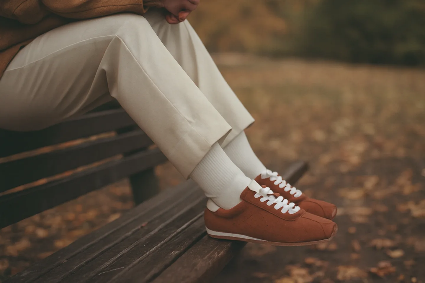 Sonbaharda parkta bir bankta oturan kadının ayakları; taba süet, ince tabanlı, beyaz bağcıklı sneaker, beyaz çorap ve bej pantolon.